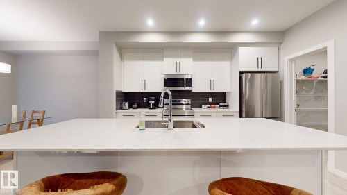 Kitchen featuring a large central island with an integrated sink and faucet, white cabinetry with dark hardware, stainless steel appliances, and a dark tile backsplash - 607 31 Avenue, Edmonton, AB 