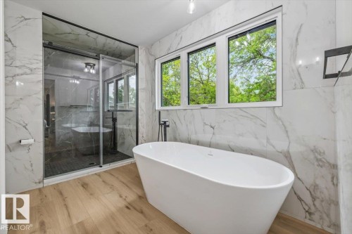 Contemporary bathroom featuring a freestanding soaking tub with a matte black floor-mounted faucet, a clear glass shower enclosure with black framing, wood-finish flooring, and large format marble-look wall tiling - 7 Glacier Place, St. Albert, AB 