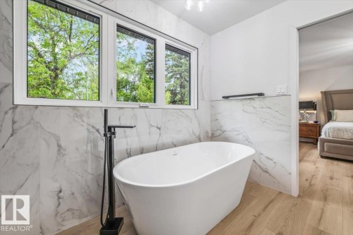 Freestanding soaking tub with a matte black floor-mounted faucet, marble-look tiled walls, light wood-finish flooring, and a large multi-pane window - 7 Glacier Place, St. Albert, AB 