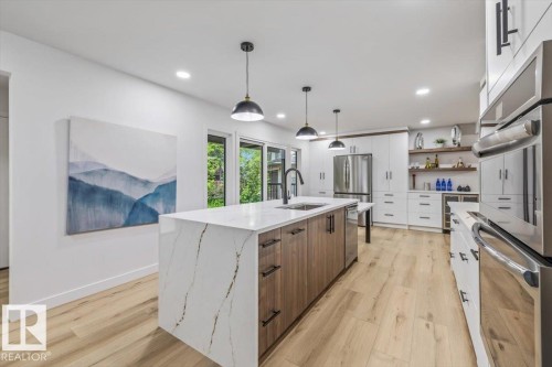 Contemporary kitchen featuring a large waterfall island with integrated sink, light wood-finish flooring, white cabinetry with dark hardware, stainless steel appliances, and built-in shelving - 7 Glacier Place, St. Albert, AB 