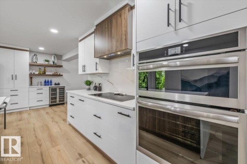 Modern kitchen featuring integrated stainless steel double ovens, white cabinetry with matte black hardware, wood-finish upper cabinets, a sleek cooktop, and light wood-finish flooring - 7 Glacier Place, St. Albert, AB 