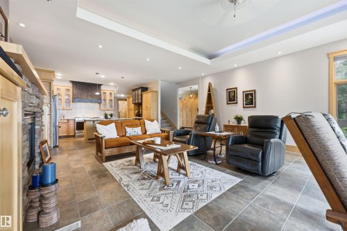 Open-concept living space featuring a stone-faced fireplace with a wood mantel, tray ceiling with recessed lighting, tile flooring, and a kitchen with stainless steel appliances and wood cabinetry - A38 Bernice Ave, Rural Leduc County, AB 