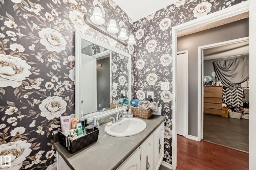 Bathroom featuring floral wallpaper, a white vanity with an integrated sink, a large framed mirror, and a three-bulb wall-mounted light fixture - 5106 19 Avenue, Edmonton, AB 