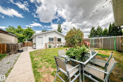 Fenced backyard with a concrete pathway and gravel border, featuring a detached structure with a white exterior, and mature trees - 5106 19 Avenue, Edmonton, AB 