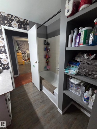 Bathroom featuring a white bathtub, gray-painted built-in shelving, wood-finish flooring, and white and black floral wallpaper - 5106 19 Avenue, Edmonton, AB 
