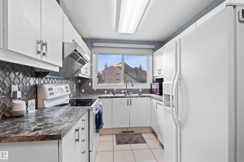 U-shaped kitchen featuring white cabinetry, dark patterned countertops, a geometric tile backsplash, and a window above the sink - 12128 25 Avenue, Edmonton, AB - Indoor Photo Showing Kitchen With Double Sink
