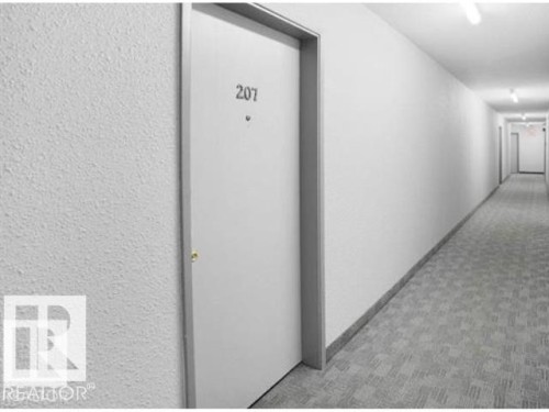 Entry door with unit number 207, set within a long corridor featuring white textured walls, recessed baseboards, and patterned carpet flooring - 107 10023 164St, Edmonton, AB 
