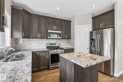 Modern kitchen featuring dark wood-finish cabinetry, stainless steel appliances, speckled countertops, a white subway tile backsplash, and wood-finish flooring - 6927 Cardinal Wynd, Edmonton, AB 
