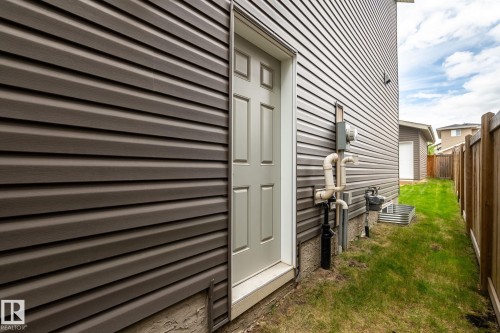 Exterior side entrance with a paneled door, dark horizontal siding, established grass, and a wooden fence - 6927 Cardinal Wynd, Edmonton, AB 