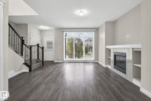 Spacious living area featuring wood-finish flooring, a contemporary fireplace with a tiled surround, built-in shelving, and a large window with vertical blinds - 6927 Cardinal Wynd, Edmonton, AB 