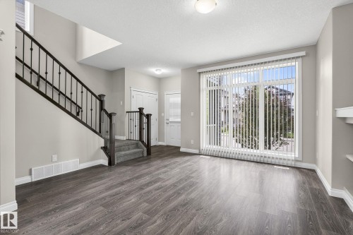 Spacious living area featuring wood-finish flooring, a contemporary staircase with wrought iron spindles, a large window with vertical blinds, and neutral wall paint - 6927 Cardinal Wynd, Edmonton, AB 