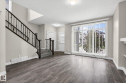 Spacious living area featuring wood-finish flooring, a contemporary staircase with wrought iron spindles, a large window with vertical blinds, and neutral wall paint - 