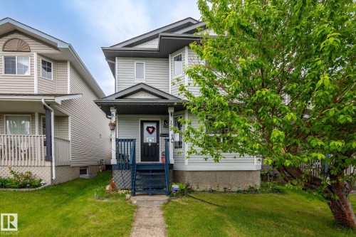 Two-story residence featuring light gray siding, a covered front porch with blue railings, a dark entry door, and a mature tree providing natural shade - 128 Brookview Wy, Stony Plain, AB 
