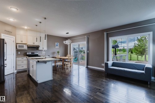 Open-concept living space featuring rich wood-finish flooring, a contemporary kitchen island with seating, white shaker cabinetry, stainless steel appliances, and a glass sliding door leading to an outdoor deck - 21223 58 Avenue, Edmonton, AB 