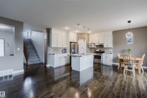 Open-concept kitchen and dining area featuring dark wood-finish flooring, white cabinetry, stainless steel appliances, a center island with a sink, and pendant lighting - 21223 58 Avenue, Edmonton, AB 