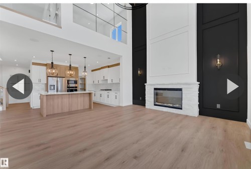 Open-concept living area featuring light wood-finish flooring, a contemporary electric fireplace with a white stone surround, and an adjacent kitchen with white cabinetry and a light wood-tone island - 13140 187 Avenue, Edmonton, AB 