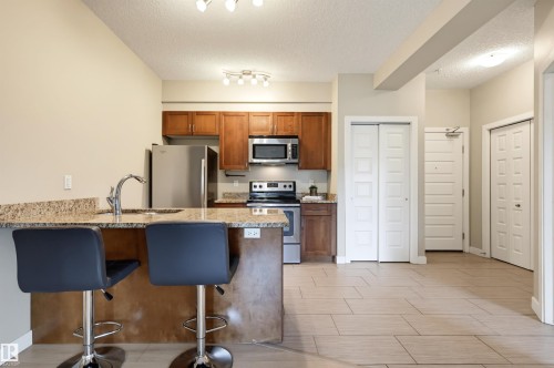 Kitchen featuring granite countertops, stainless steel appliances, wood cabinetry, and a breakfast bar - 115 1238 Windermere Way, Edmonton, AB 