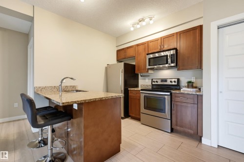 Modern kitchen featuring stainless steel appliances, granite countertops, wood cabinetry, and a peninsula with a built-in sink - 115 1238 Windermere Way, Edmonton, AB 