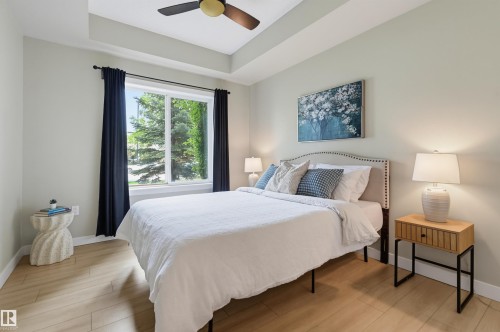 Recessed tray ceiling with a ceiling fan, large window with dark curtain panels, wood-finish flooring, and light neutral wall paint - 115 1238 Windermere Way, Edmonton, AB 