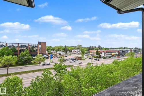 Urban landscape featuring a prominent commercial building with a glass facade and brick accents, surrounded by mature trees - 402 8730 82 Avenue, Edmonton, AB 