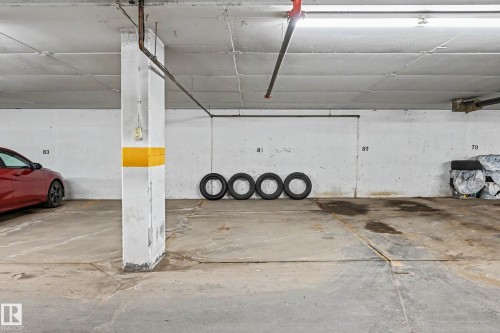 Designated parking space within a concrete garage structure, featuring a painted concrete floor, white walls, overhead fluorescent lighting, and visible fire suppression piping - 402 8730 82 Avenue, Edmonton, AB 