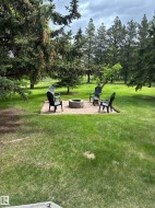 Expansive backyard featuring a central paver patio with a built-in stone fire pit, surrounded by mature evergreen trees - 
