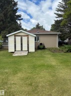 Expansive grassy backyard featuring a storage shed with double doors and a wood ramp, a stucco-sided exterior, a brown shingle roof, and mature perimeter trees - 