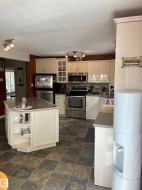 Spacious kitchen featuring an angled island with built-in shelving, durable stone-finish tile flooring, recessed ceiling lighting, cream-colored cabinetry, and stainless steel appliances - 