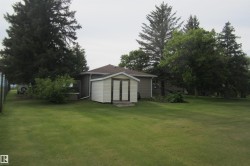 Expansive lawn area featuring a detached storage shed with double doors, mature evergreen trees, deciduous trees, and a partial fence line - 