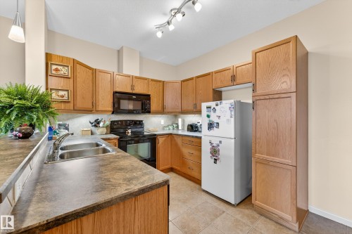 Kitchen featuring wood cabinetry, a double basin stainless steel sink, black appliances, and tiled flooring - 133 2503 Hanna Crescent, Edmonton, AB 