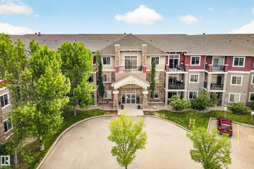 Multi-story residential building featuring stone and siding exterior accents, a prominent covered entrance, private balconies, a circular driveway, and mature landscaping - 133 2503 Hanna Crescent, Edmonton, AB 