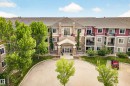 Multi-story residential building featuring stone and siding exterior accents, a prominent covered entrance, private balconies, a circular driveway, and mature landscaping - 133 2503 Hanna Crescent, Edmonton, AB 