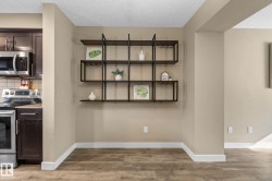 Open concept area featuring wood-finish flooring, a contemporary wall-mounted shelving unit with integrated stemware racks, and a kitchen with stainless steel appliances, dark cabinetry, and a subway tile backsplash - 