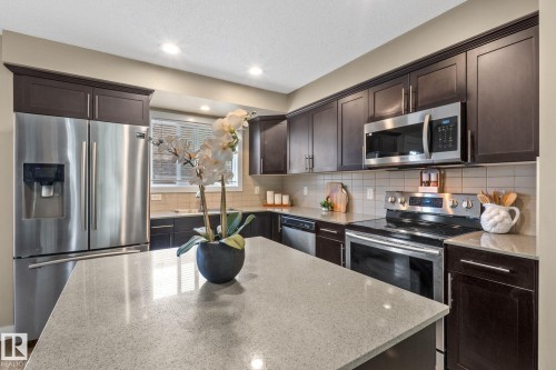 Kitchen featuring dark wood-finish cabinetry, stainless steel appliances, a light-toned tile backsplash, and a spacious island with a light-speckled countertop - 12809 203A, Edmonton, AB 