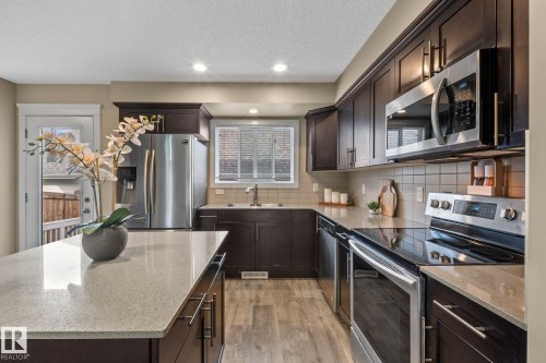 Contemporary kitchen featuring dark wood cabinetry, light-toned countertops, stainless steel appliances, a tiled backsplash, and wood-finish flooring - 12809 203A, Edmonton, AB 