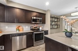 Modern kitchen featuring dark wood-finish cabinetry, stainless steel appliances, light-toned countertops, a light-colored tile backsplash, and wood-finish flooring - 