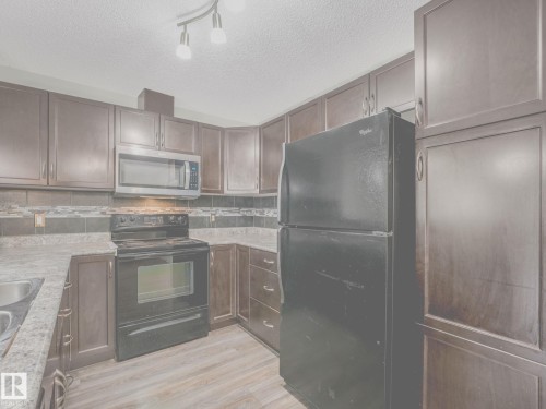 Kitchen featuring dark wood cabinetry, a stainless steel microwave, black electric range, a black refrigerator, and wood-finish flooring - 117 6070 Schonsee Way, Edmonton, AB 