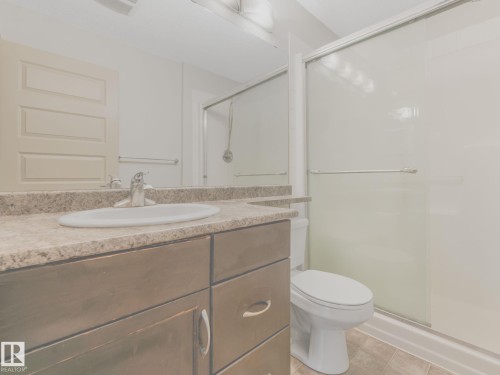 Bathroom featuring a dark wood-finish vanity with an integrated sink, a neutral-toned countertop, a shower with a sliding glass door, and tiled flooring - 117 6070 Schonsee Way, Edmonton, AB 