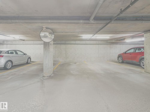 Underground parking garage featuring concrete walls and ceiling, painted concrete flooring, overhead fluorescent lighting, and a convex mirror for visibility - 117 6070 Schonsee Way, Edmonton, AB 