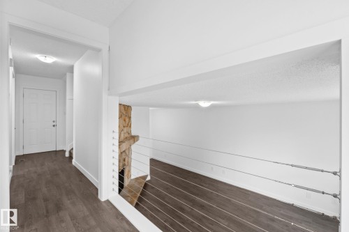 Upper-level walkway featuring wood-finish flooring, a white paneled door, and a modern cable railing system overlooking a lower living space with a stone fireplace - 2832 135 Avenue, Edmonton, AB - Indoor Photo Showing Other Room