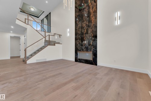 Two-story living area featuring a prominent dark marble-look fireplace with an integrated hearth, light wood-finish flooring, and a contemporary open-riser staircase with glass railings and wood handrails - 3602 44 Avenue, Beaumont, AB 