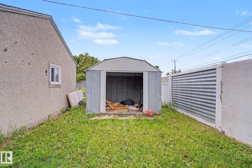 Rear yard featuring a standalone storage shed, a stucco exterior wall with a white-framed window, and a corrugated metal fence panel - 11914 124 Street, Edmonton, AB 