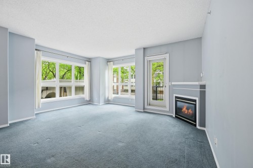 Living area featuring an integrated fireplace with a corner mantel, light blue carpeting, multiple windows, and a glass-paneled door - 203 11103 84 Avenue, Edmonton, AB 