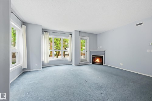 Living area featuring a corner-set fireplace with a white mantel, light blue carpeting, and multiple windows - 203 11103 84 Avenue, Edmonton, AB 