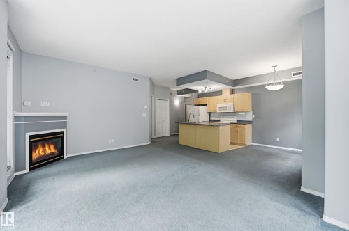 Open-concept living area featuring a built-in fireplace, light wood cabinetry, a kitchen island with countertop seating, and a pendant light fixture - 203 11103 84 Avenue, Edmonton, AB 