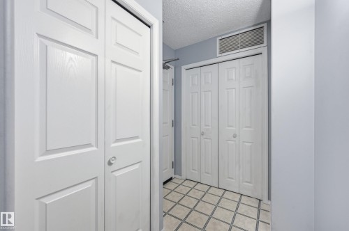 Hallway featuring panel-style bifold closet doors, light blue wall paint, and square tile flooring with contrasting grout - 203 11103 84 Avenue, Edmonton, AB 