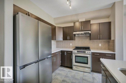 Modern kitchen featuring stainless steel appliances, dark wood-finish cabinetry, light-toned countertops, tile backsplash, and track lighting - 14 4367 Veterans Way, Edmonton, AB 