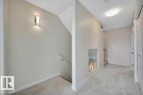 Carpeted hallway featuring a built-in wall niche with glass paneling, overhead flush mount lighting, and contrasting baseboards - 14 4367 Veterans Way, Edmonton, AB 