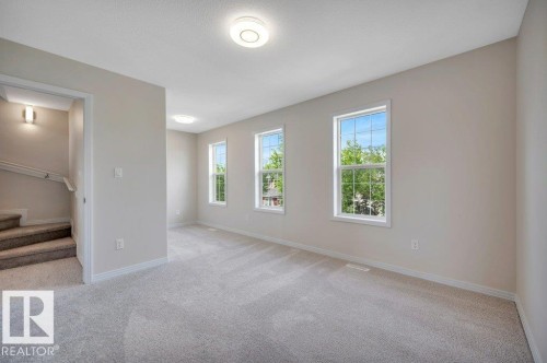 Carpeted open-concept room featuring three windows, a ceiling-mounted light fixture, and neutral wall paint - 14 4367 Veterans Way, Edmonton, AB 