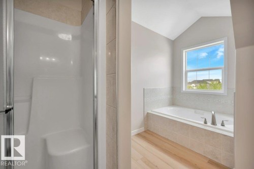 Bathroom featuring a built-in shower with a seat, a soaking tub with tile surround, a large window, and wood-finish flooring - 14 4367 Veterans Way, Edmonton, AB 